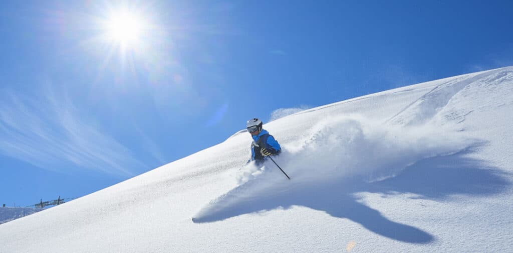 Skier descending a snow-covered mountain under bright sunlight at high altitude.