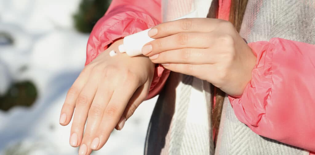 Woman applying skincare cream to her hand outdoors in snowy winter conditions.
